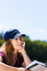 An outdoor portrait of a young woman wearing a blue cap, reading a book in a tranquil setting,...