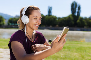 A happy young woman wearing headphones listens to music and uses a smartphone in a sunny park by...