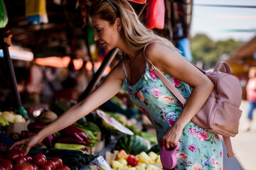 A stylish young woman reaches for tomatoes at a vibrant outdoor market, selecting fresh produce on...