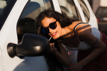 Charming young woman in sunglasses using a car mirror to apply lipstick during a radiant sunny day,...