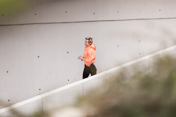 A fit young woman wearing a fluorescent orange jacket jogs down a concrete ramp, depicting an...