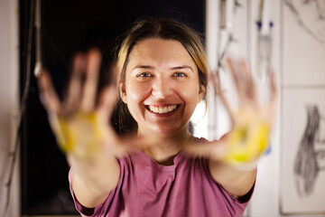 Smiling woman artist with colorful paint on her hands standing in her art studio, displaying a...