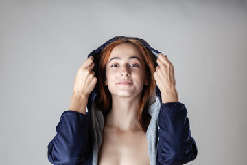 A young female model gently pulls up the hood of her dark blue jacket, revealing a playful expression and bare shoulders against a neutral gray backdrop.