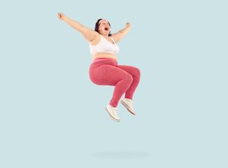 Full length shot of funny chubby overweight woman jumping in air. Happy plus size, fat woman wearing sportswear jumping high with her hands outstretched isolated on light blue studio background