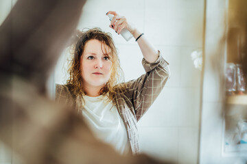 Haircare. Young plus size Lady Applying Hair Spray For Curls While Standing Near Mirror In Bathroom, Woman takes care of wavy hair with sea salt spray. Haircare cosmetics and beauty routine concept.