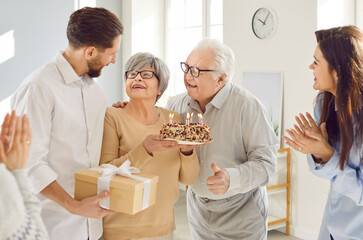 Elderly father and adult children celebrate elderly mother at home with a birthday cake and gifts. The family gathers for a joyous party, marking this special anniversary with happiness and love.