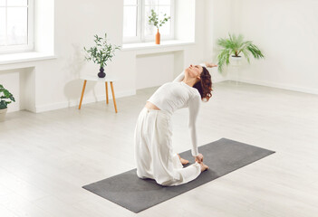 Young yogi sporty attractive woman practicing yoga, sitting in Ustrasana exercise, Camel pose, working out, wearing white sportswear, home interior background, near potted plants