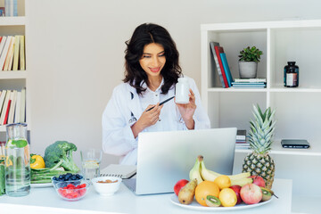 Positive Indian female nutritionist having online consultation with client at office desk with fresh vegetables, prescribing nutritional supplements and vitamins, making balanced corrective diet plan.