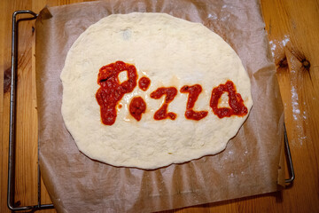Unbaked pizza dough on parchment paper with tomato sauce spelling “pizza” in a cozy kitchen setting.