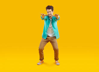 Cheerful funny guy jokingly pretending to squat with arms outstretched, isolated on orange background. Young Caucasian man in casual shirt, t-shirt and pants is fooling around and smiling. Full length
