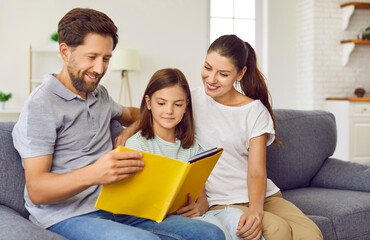 Young happy family reading a book with their little cute daughter sitting on sofa at home. Smiling...