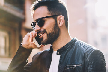 Cheerful hipster guy happy to hear friend making mobile call in roaming satisfied with connection,...