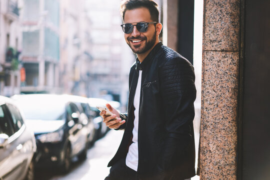 Half length portrait of cheerful hipster guy using smartphone standing on city street blogging in social networks,happy handsome male looking at camera chatting with friend online via telephone