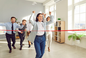 Happy excited woman employee running to red ribbon celebrating crossing finishing line in office with people colleagues in background. Business competition, success and teamwork concept.