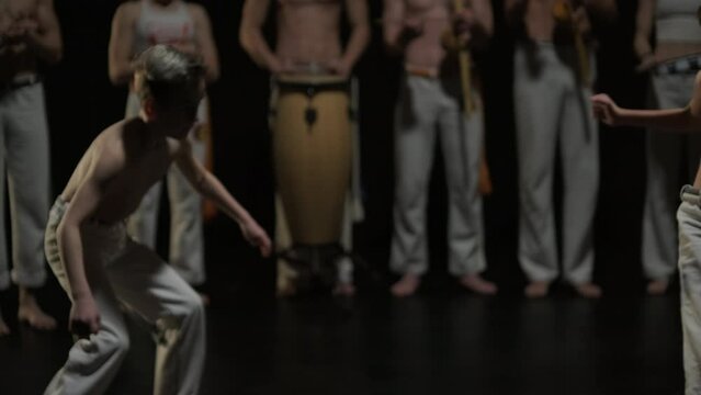 Group of a professional dancers are practicing capoeira in darkness against a spotlight on a black background of studio. Afro-Brazilian martial art that combine elements of dance.