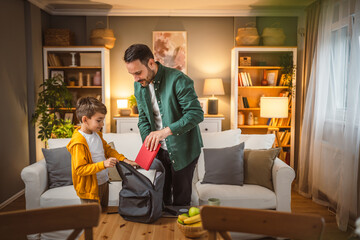 Naklejka premium Dad help his son to pack his notebook and book for school in backpack