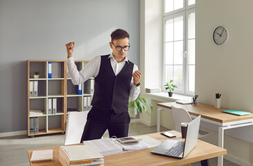 Happy emotional confident accountant standing by office desk with computer raising fists up. Man...