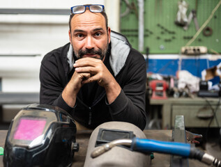 Nice portrait of a Caucasian man looking into camera. On his bench are his welder's work tools: two protective helmets and the welding gun.