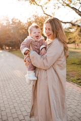 Smiling woman 25-29 year old wearing beige trench coat holding baby girl over yellow leaves outdoor in park. Autumn season. Motherhood.