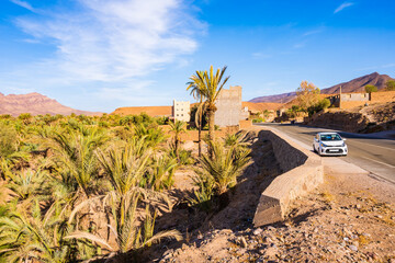 Rental car on scenic road in green palm tree oasis of Agdz town with mountains in background,...