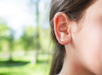 A young woman displays a stylized fleur-de-lis gold earring on her right ear in a natural outdoor...