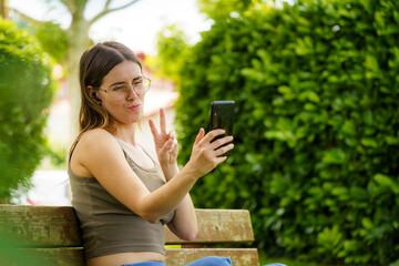 Young Caucasian Girl Looking at Her Phone and Making a Peace Gesture. Sitting on a Bench in an...