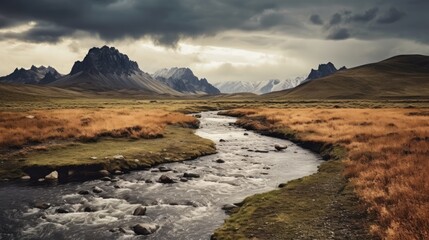 Obraz premium Dramatic mountain landscape with a winding river under moody skies in a vast, grassy plain with snow capped peaks in the distance