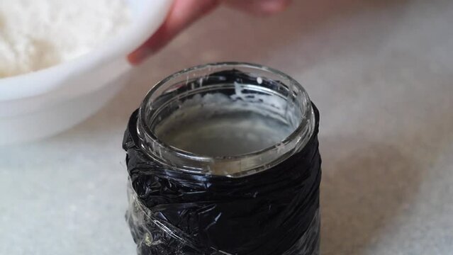 Video of pouring flour with a tablespoon into a glass jar wrapped in a black bag to obtain yeast-free sourdough for further baking homemade bread. An old traditional recipe