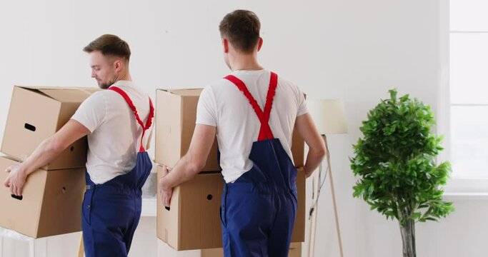 Young delivery men carrying cardboard boxes, movers service unloading and removing things. Moving company working, workmen wearing uniform helping to change residence packing, entering new flat 