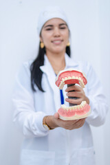In her office, a dental model stands out in the foreground while in the background there is a Latina dentist with a big smile, wearing a waterproof uniform, gown, and cap.