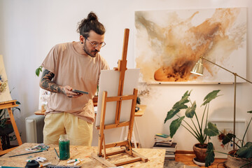 A creative tattooed artist painting an artwork on easel at atelier.
