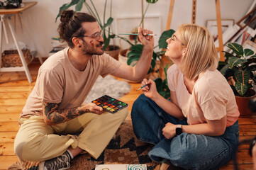Artistic couple sitting on the floor at studio and painting on face.