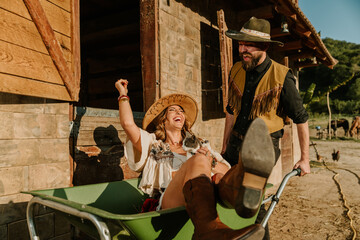 Young happy farmer couple having fun with wheelbarrow at ranch.