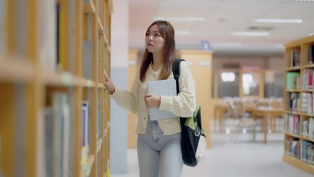 A young woman is browsing through a library, looking for a book. She is holding a book in her hand and has a backpack on her back. The library is filled with bookshelves