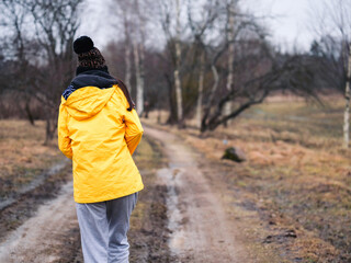 A teenager girl in a yellow jacket is walking down a muddy road. Concept of solitude and...