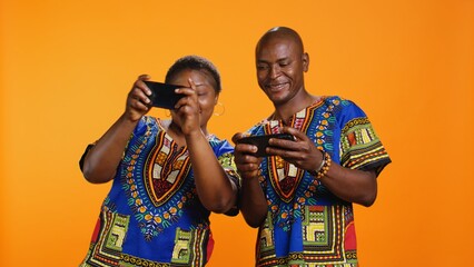 Obraz premium African american people playing videogames on mobile device, having fun with online gaming contest in studio. Married man and woman enjoying game tournament on smartphone app.