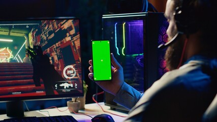 Man watching tutorial on green screen phone while playing first person shooter videogame with gun...