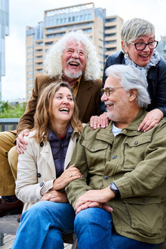 Vertical. Two couple of elderly Caucasians laughing together sitting on bench in city. Happy old friends gathered having fun outdoors. Lifestyle of cheerful mature people enjoying sunny autumn day