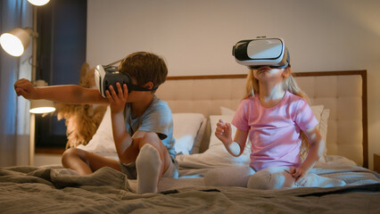 Curious modern little Caucasian children kids wearing virtual reality headset vr helmet glasses son boy interacting digital 3d world playing game wireless technology girl daughter midnight bed bedroom