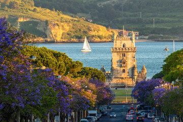 Belem Tower, Jacaranda Blooming Purple Blue Trees and Sailboat on Sunny Evening. Golden Hour. Lisbon, Portugal. © maxterram