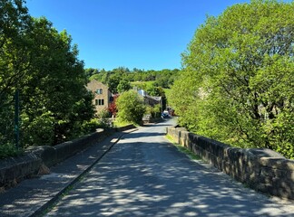 On a sunny day, shadows are cast by an ancient stone bridge that leads to a group of buildings nestled among verdant trees in Kilnhurst, Todmorden, Yorkshire, UK.