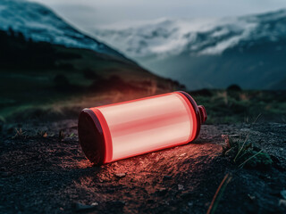 A glowing red battery lying on a rock in a mountainous landscape during twilight. Snow-capped mountains and a valley are visible in the background.