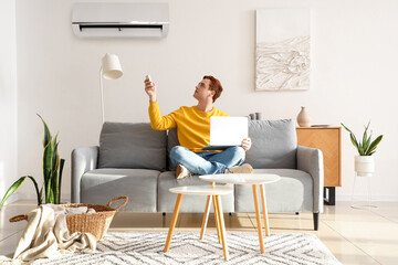 Young man with laptop turning on air conditioner on couch in living room