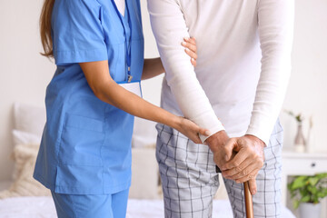 Female caregiver helping senior man with walking stick in bedroom