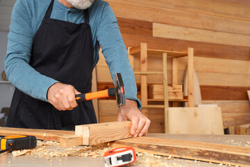 Mature carpenter hammering nail into wooden plank at table in shop, closeup
