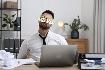 Man with fake eyes painted on sticky notes at workplace in office. Overwhelmed by work