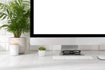 Office workplace with computer, glasses and houseplants on light table near white brick wall
