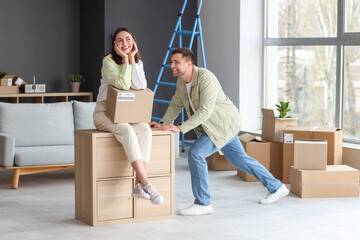 Happy young couple with box and chest of drawers in room on moving day