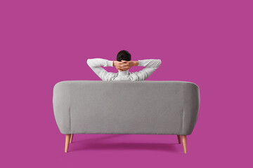Handsome young man resting on sofa against purple background, back view