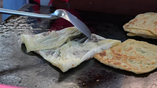 Street food vendor is preparing the dough and pan-frying paratha roti in hot oil until crispy on the outside. Street hawker preparing prata bread or roti canai for morning breakfast
